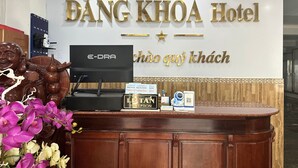 Bancada de recepção do Hotel ĐÀNG KHÔA, com televisão, placa indicativa da recepção e uma estátua decorativa.