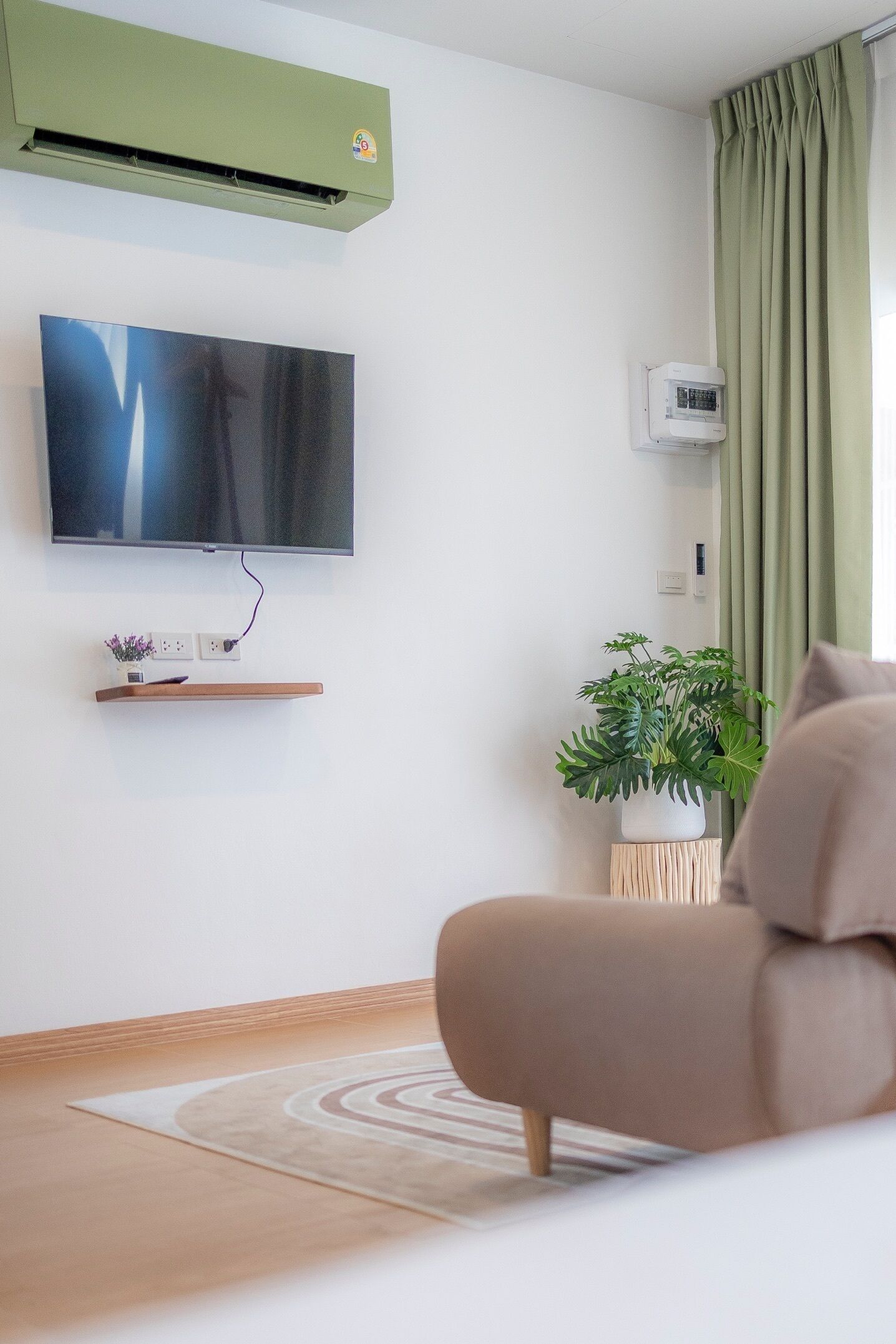 A modern living room with a flat-screen TV mounted on the wall, a green air conditioner, a potted plant, and a beige sofa.
