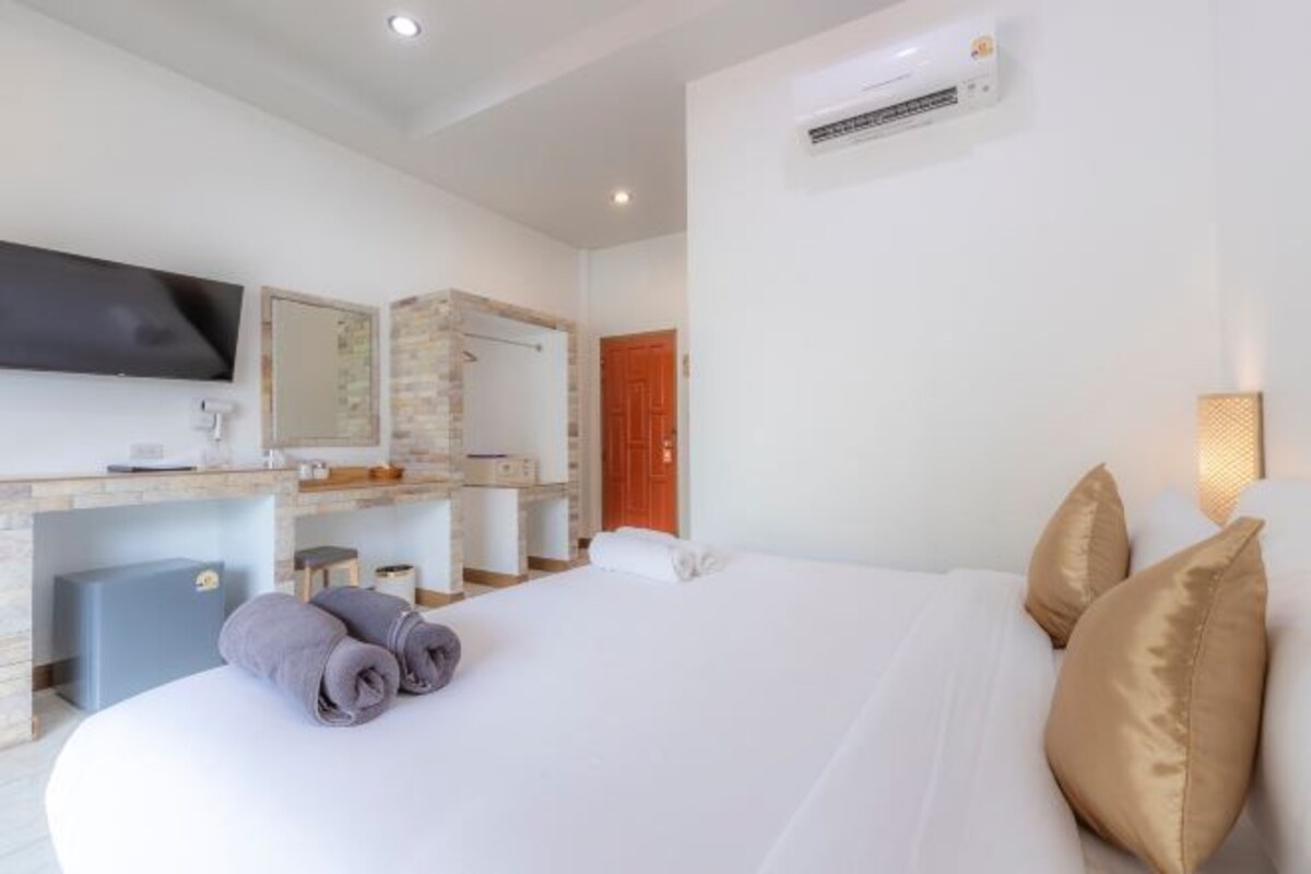 A hotel room with a large bed, a flat-screen TV mounted on the wall, a desk with a computer, and a bathroom with a shower and toilet.