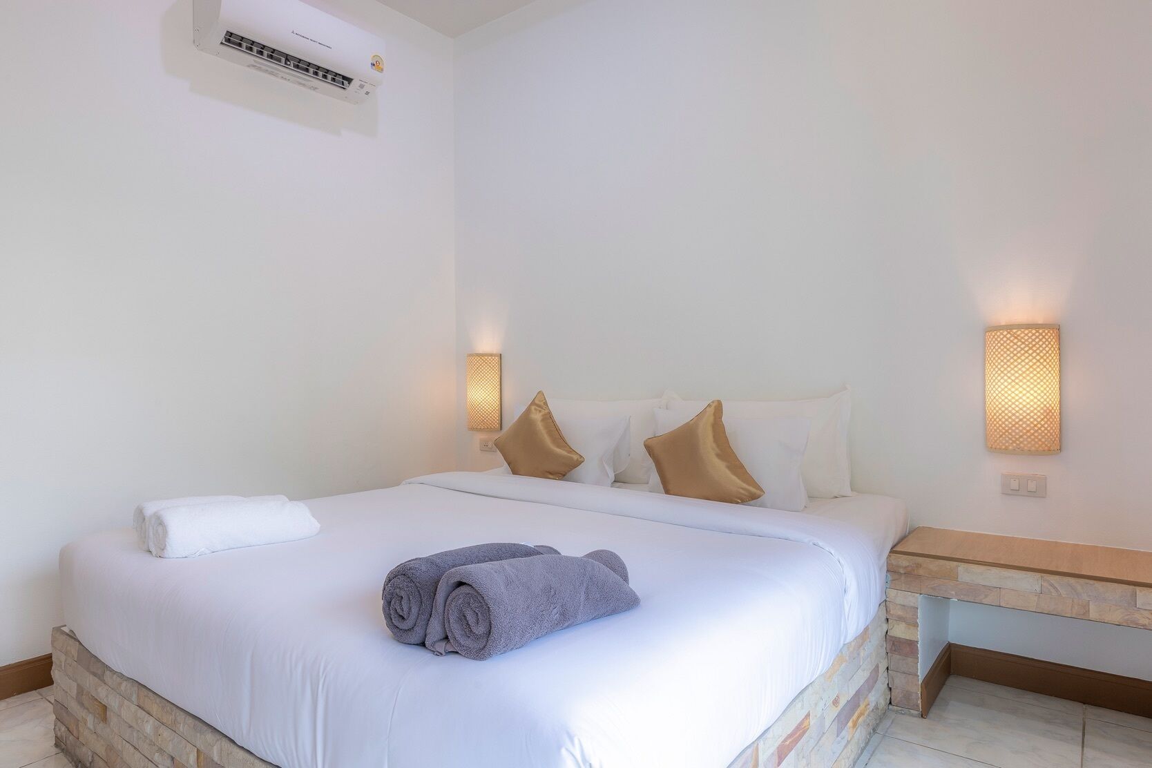 A hotel room with a bed, white linens, a rolled towel, two wall-mounted lamps, and an air conditioning unit.