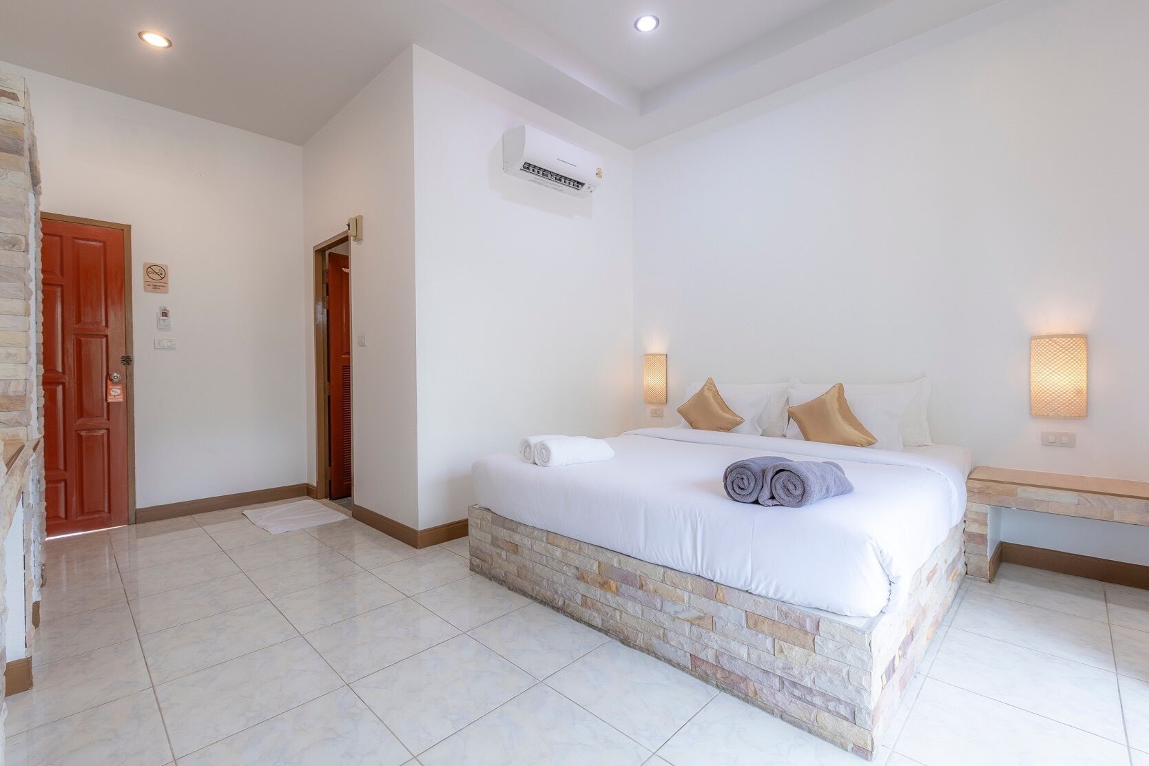 A hotel room with a bed, a nightstand, a wall-mounted air conditioner, and a door with a sign.