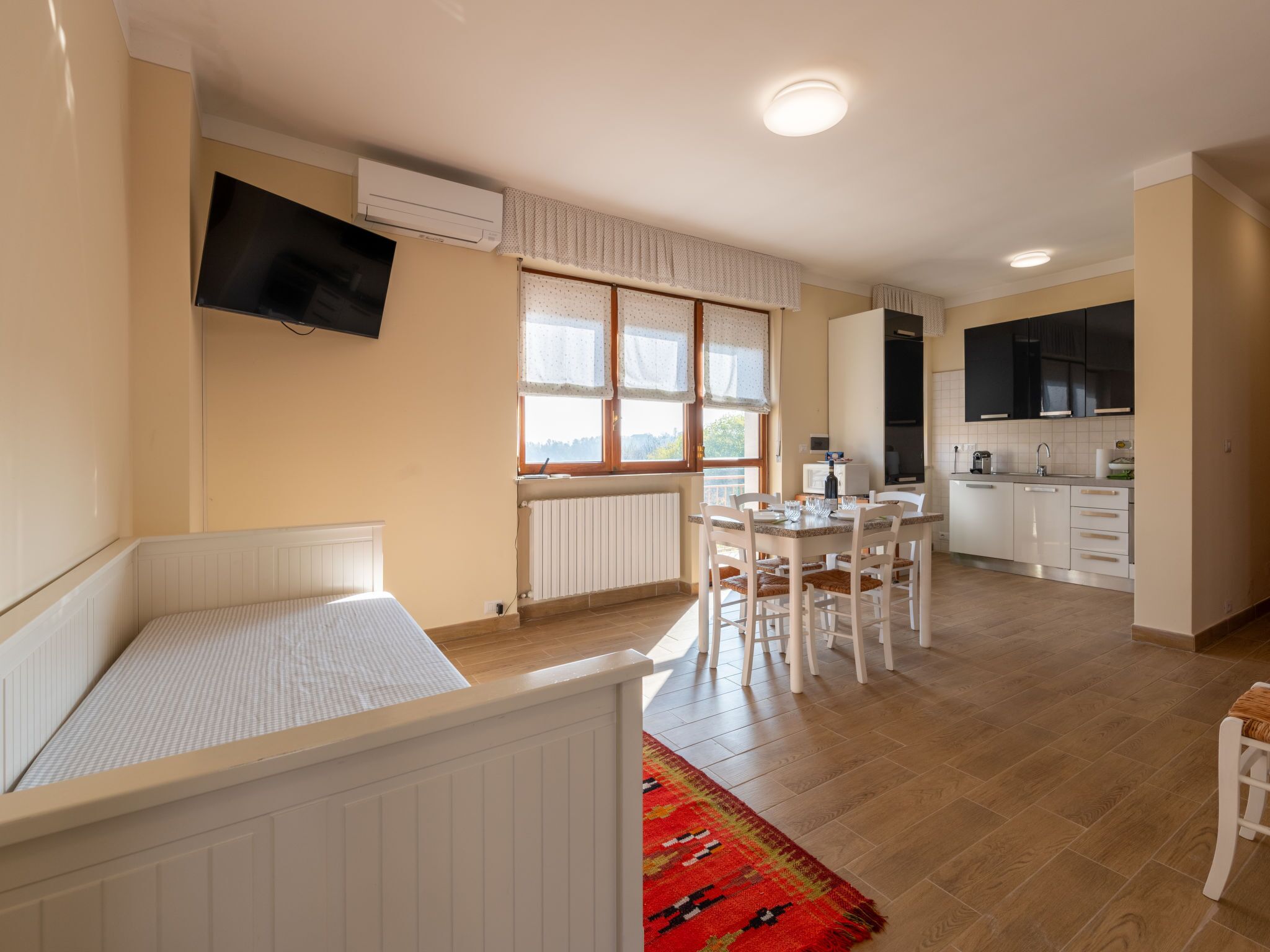 A modern, well-lit room with a bed, a dining table with chairs, a kitchen area, and a TV mounted on the wall.