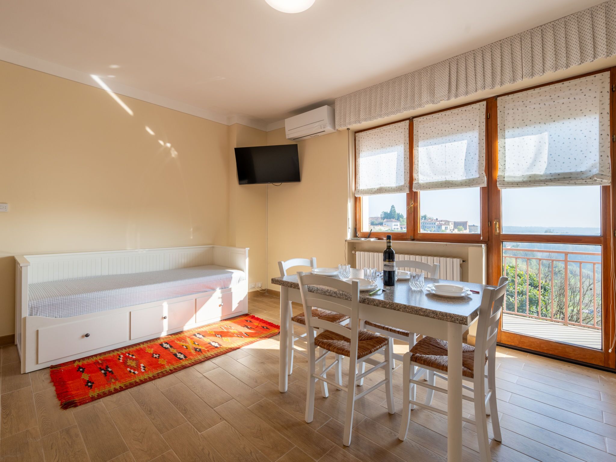 A room with a dining table, chairs, a bench, a TV mounted on the wall, and a window with a view.
