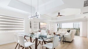 Un vaste espace de vie comprenant une table à manger en verre, des chaises blanches, un canapé et un ventilateur de plafond.