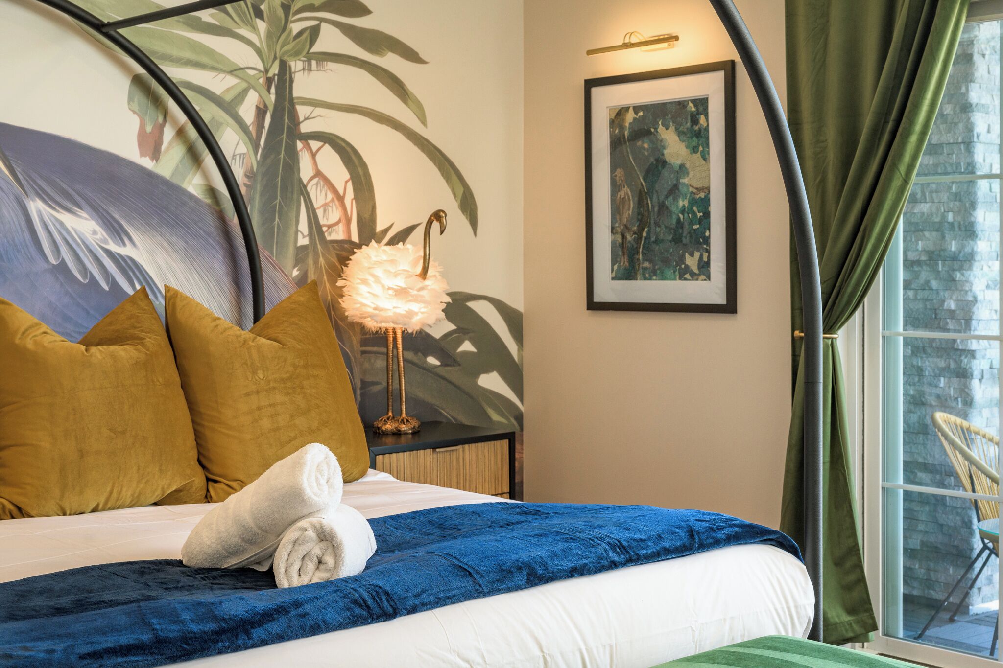 Een netjes opgemaakt bed met een blauwe deken, een witte handdoek en twee mosterdgele kussens. Aan de muur hangt een ingelijst schilderij en op een nachtkastje staat een lamp.
