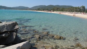 Uma praia com águas claras, pedras em primeiro plano e uma faixa de areia com guarda-sóis e pessoas.
