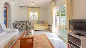 Una sala de estar con sofá, una mesa de centro, mueble para televisión, una mesa de comedor con sillas, una ventana con cortinas y una puerta corrediza de vidrio.
