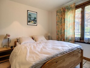 A bedroom with a bed, two bedside tables, a nightstand, a lamp, a painting on the wall, and a window with curtains.