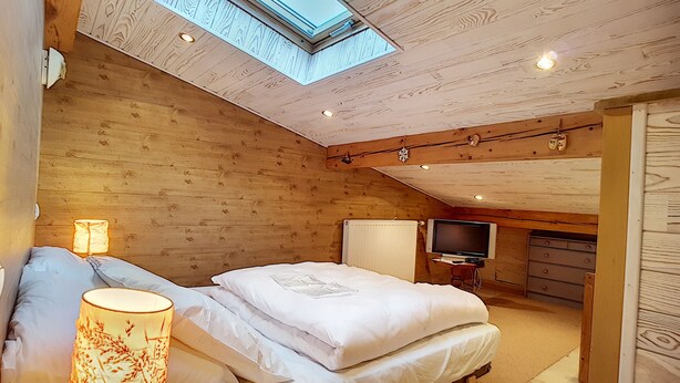 A cozy wooden cabin bedroom with a skylight, a bed with white linens, a bedside lamp, and a television on a stand. - Clos 28 - appartement - 6pers - proche centre (Les Gets)