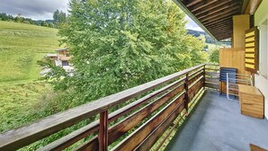 A balcony with a wooden railing overlooking a green landscape and a building. - Virola - appartement - 4 pers - proche piste (Les Gets)