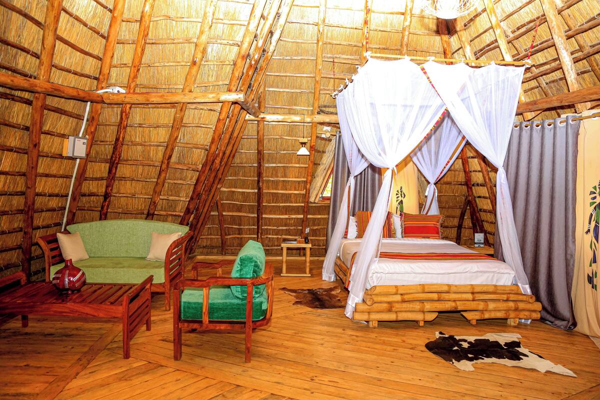 A thatched-roof room with a bed, a green sofa, a wooden chair, and a small table.