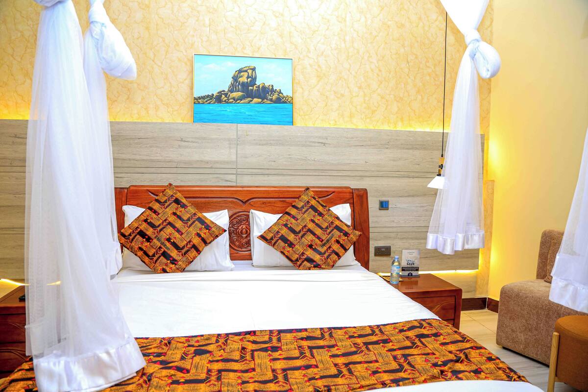 A hotel room with a large bed, a wooden headboard, two patterned pillows, a painting on the wall, and a chair.