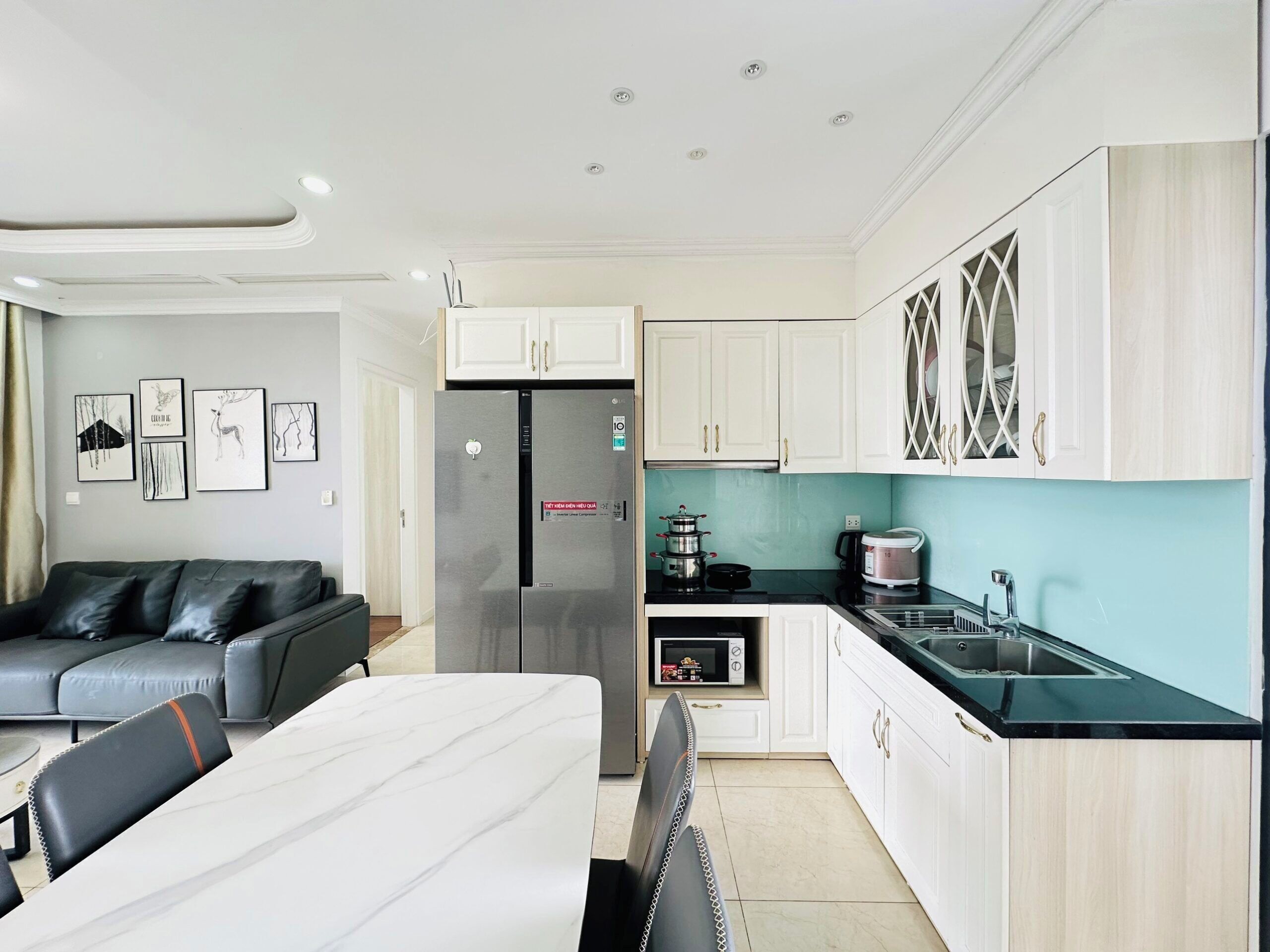 A modern kitchen with white cabinets, a stainless steel refrigerator, a microwave, and a sink. There is a dining table with chairs and a grey sofa in the living area.