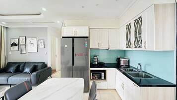 A modern kitchen with white cabinets, a stainless steel refrigerator, a microwave, and a sink. There is a dining table with chairs and a grey sofa in the living area.
