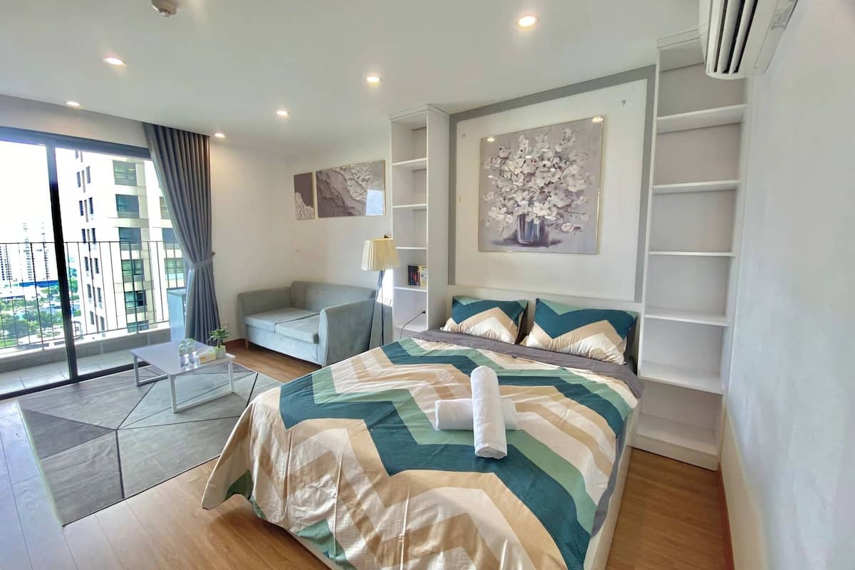 A modern bedroom with a large bed, a sofa, a small table, and a view of the city through large windows.