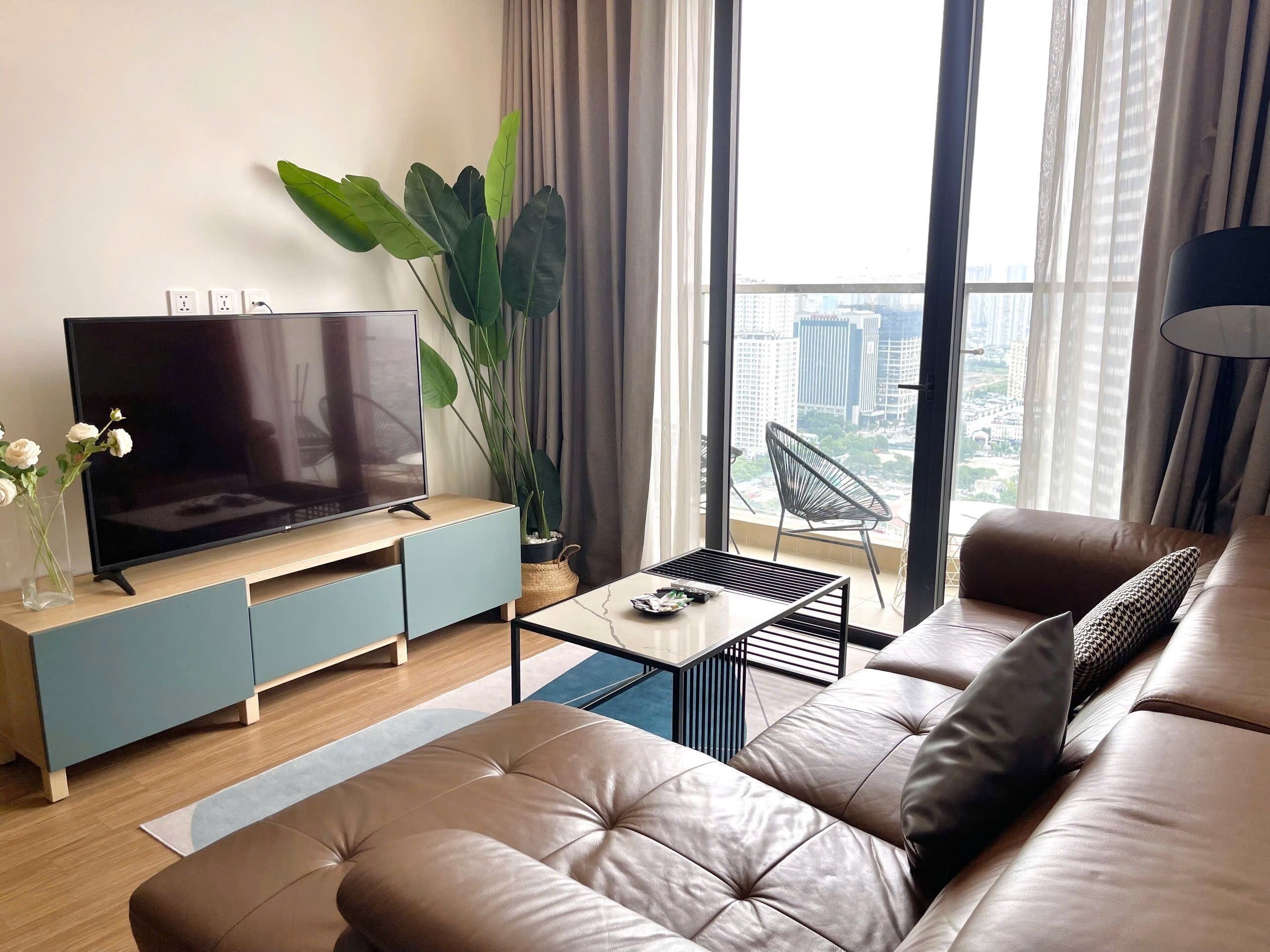 A modern living room with a flat-screen TV on a wooden TV stand, a large potted plant, a comfortable sofa, and a view of the city through large windows.