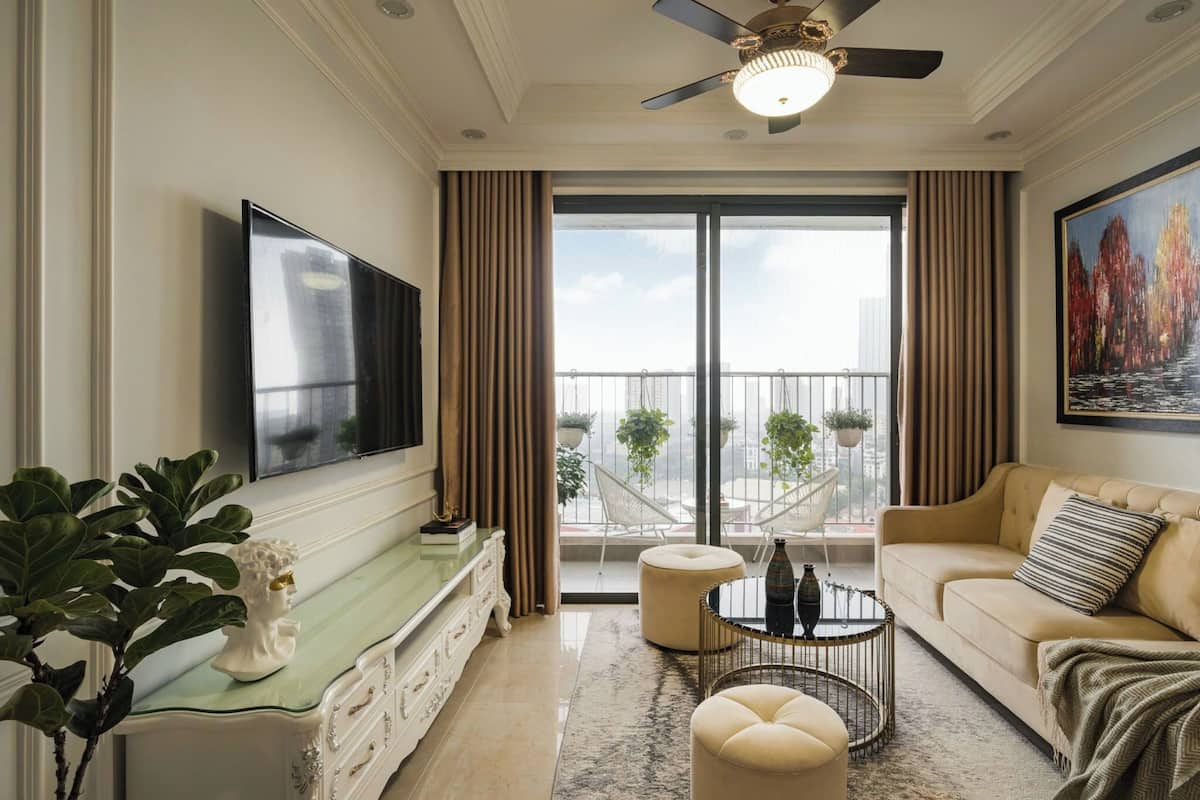 A spacious living room with a large flat-screen TV, a beige sofa, a coffee table, and a balcony with potted plants.