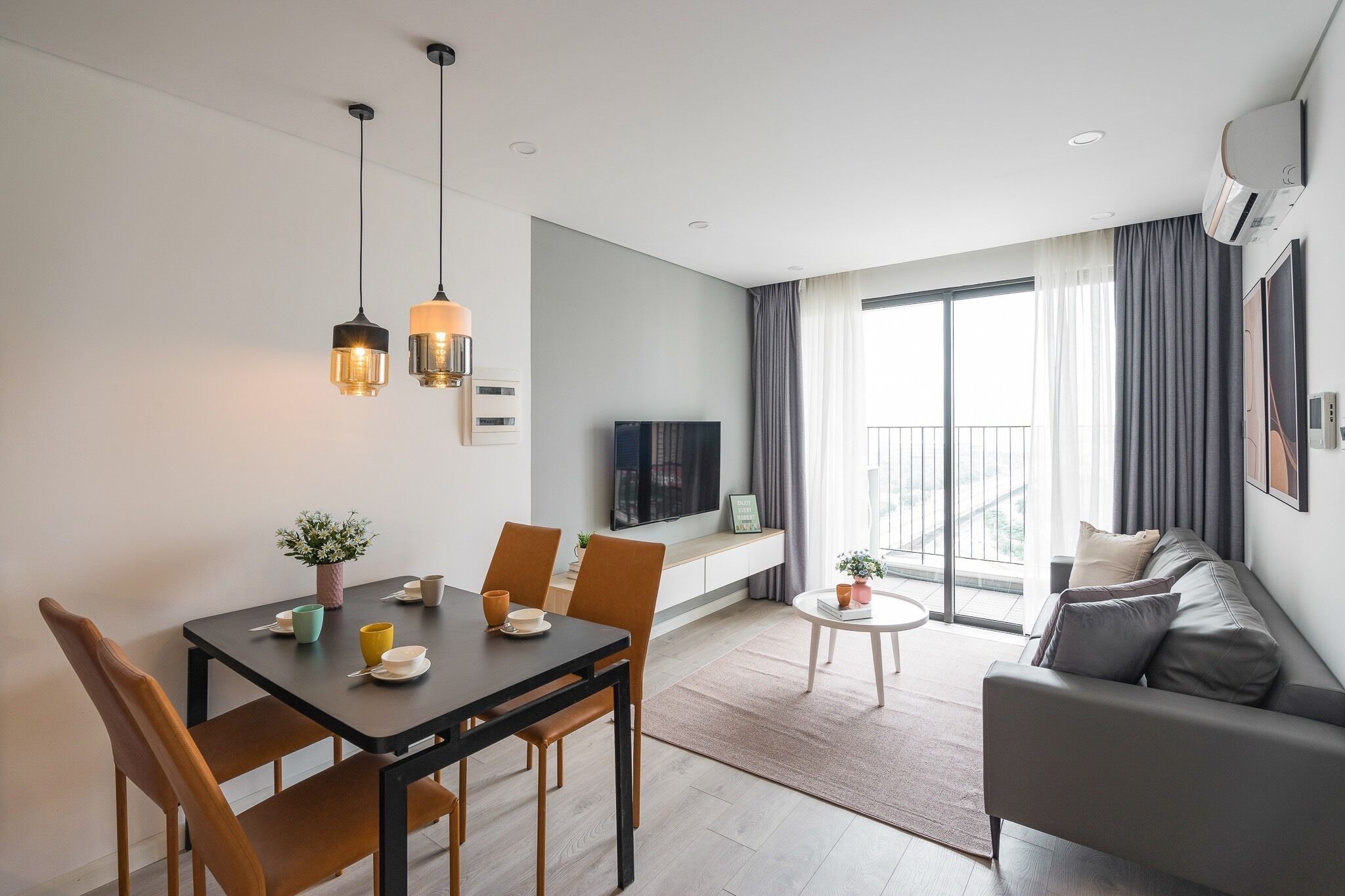 Ein modernes Wohnzimmer mit einer Couch, einem Esstisch mit Stühlen, einem an der Wand befestigten Fernseher und einem Balkon mit Vorhängen.