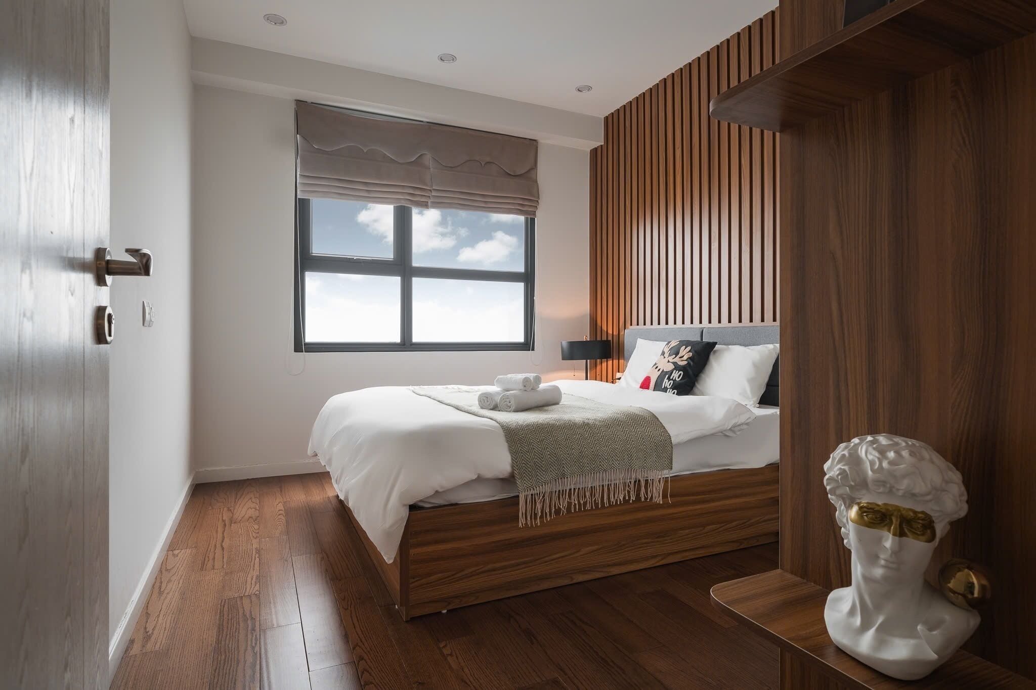 Een moderne slaapkamer met een houten bed, een groot raam met uitzicht en een decoratief buste op een nachtkastje.
