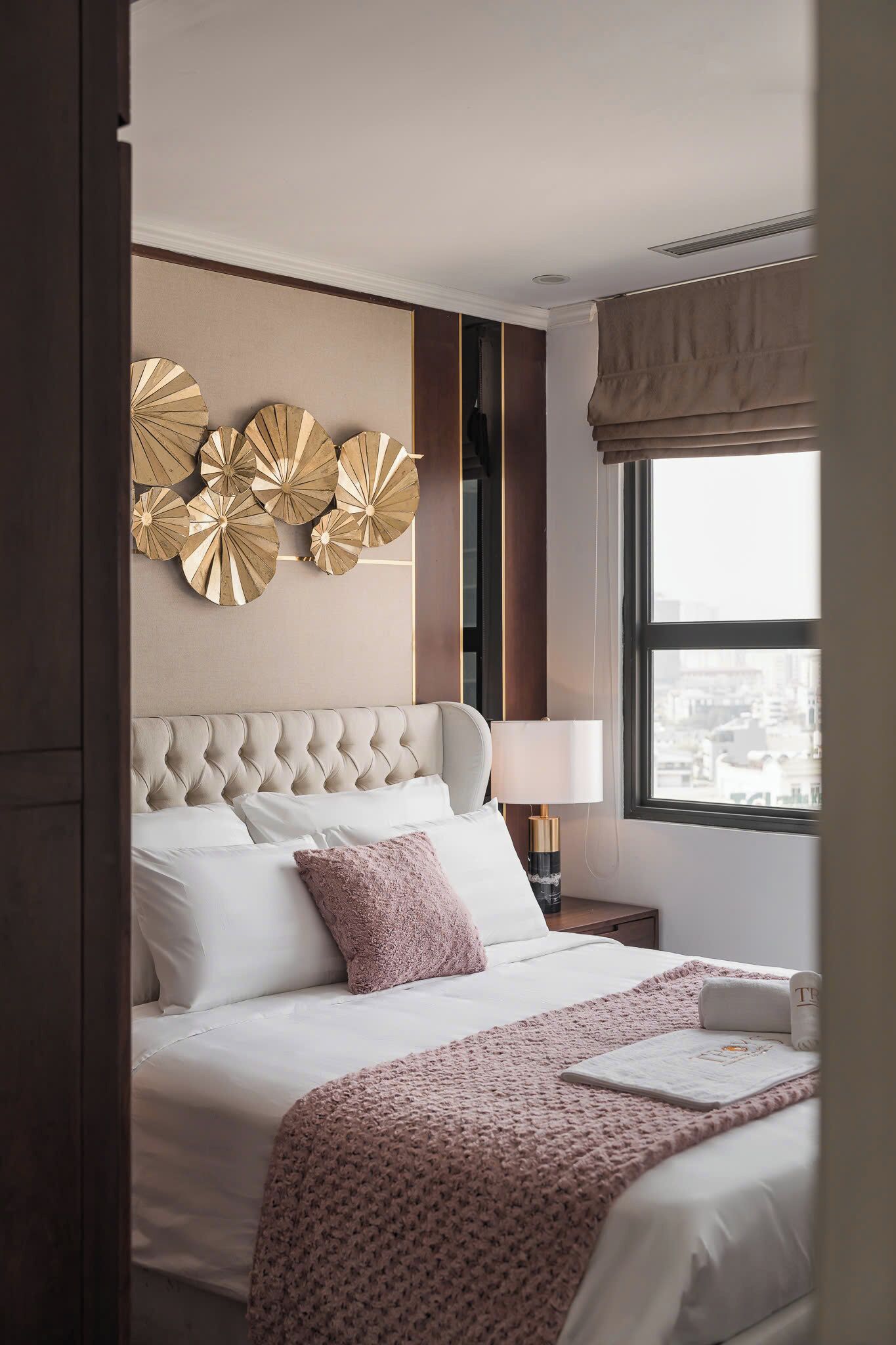 A hotel room with a tufted headboard, a large window with a city view, a decorative wall piece, and a bedside table with a lamp.