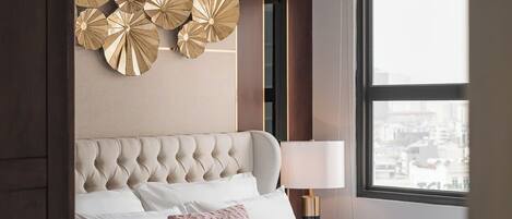 A hotel room with a tufted headboard, a large window with a city view, a decorative wall piece, and a bedside table with a lamp.