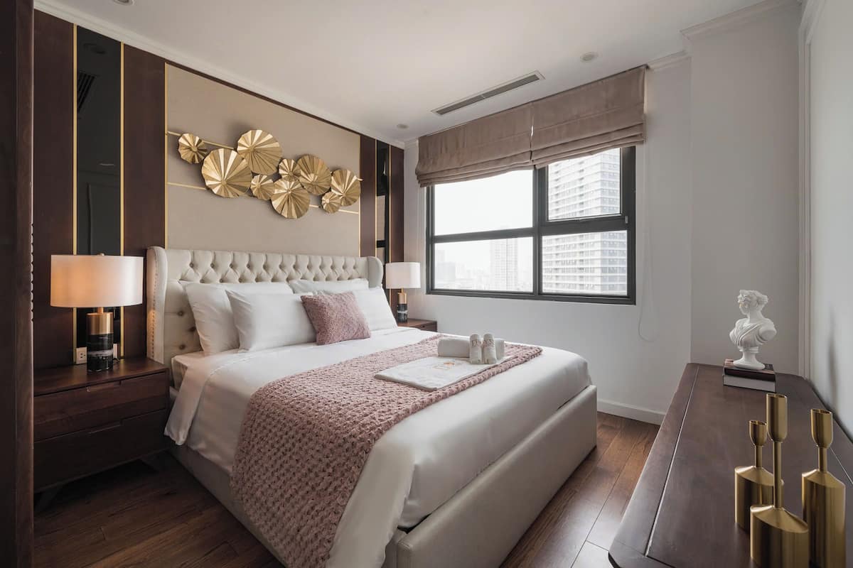 A modern hotel room with a large bed, a bedside table with a lamp, a decorative wall piece, and a view of the city through the window.