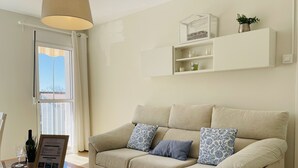 A living room with a beige sofa, a glass coffee table, a white throw blanket, and a mounted shelf with decorative items.