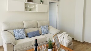 A living room with a beige sofa, patterned cushions, a glass coffee table, a wine bottle, glasses, a potted plant, and a framed picture.
