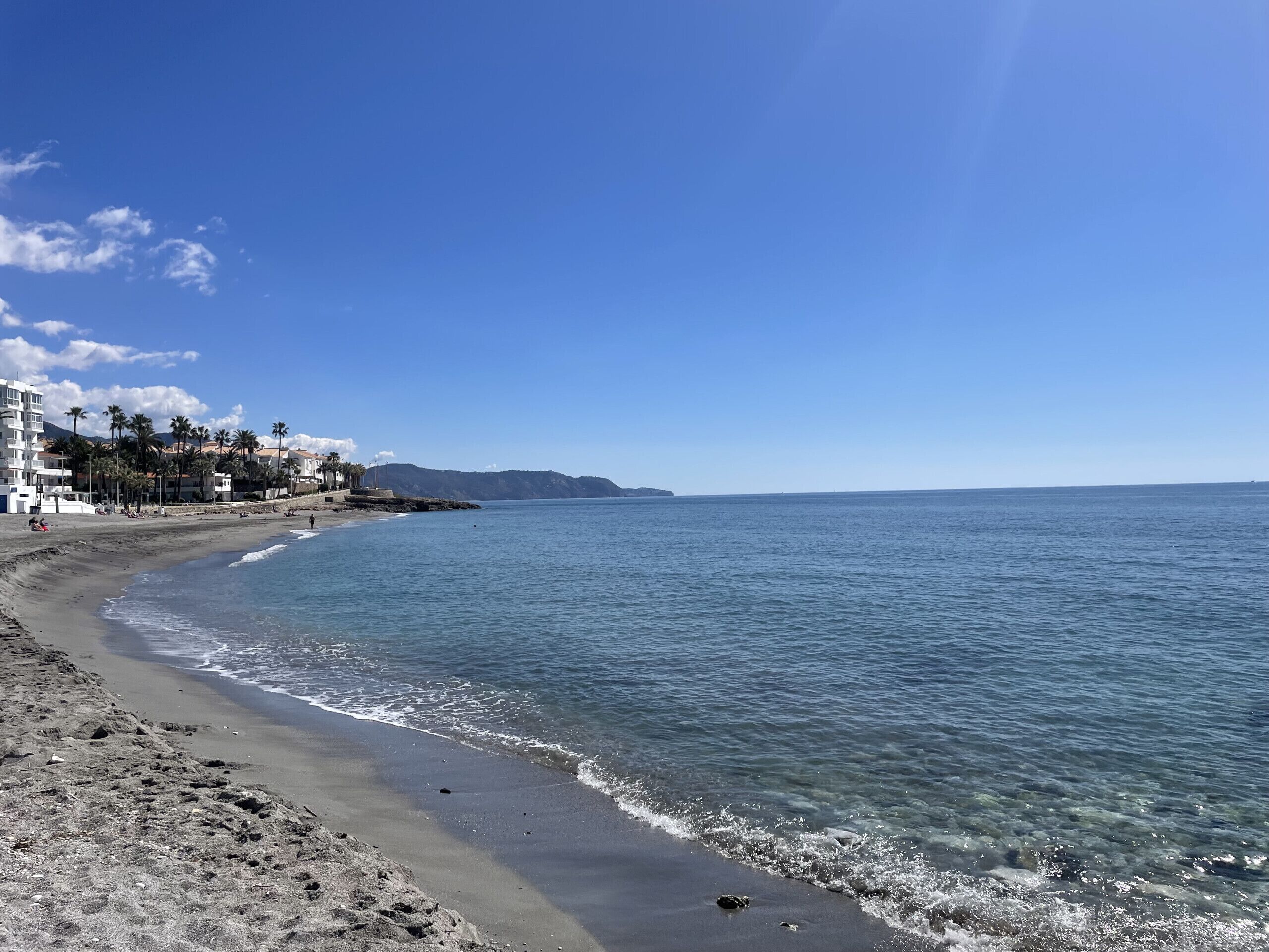 A beach with clear blue water, a sandy shore, and buildings in the background.