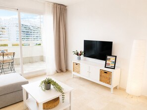 A modern living room with a flat-screen TV, a sofa, a dining table, and a balcony with a view of the city.