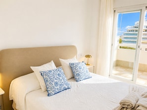 A hotel room with a bed, two pillows, a nightstand with a lamp, and a view of a building through a glass door.