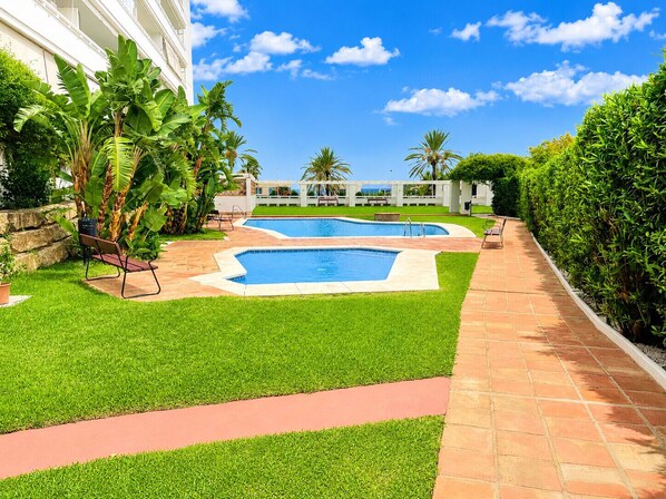 A swimming pool area with two pools, surrounded by well-maintained grass and a brick pathway.
