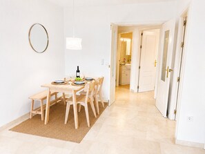A dining area with a table set for four, a bottle of wine, and a fruit bowl. There are two chairs and a bench. The room has white walls, a hanging light fixture, and a door leading to a bathroom.