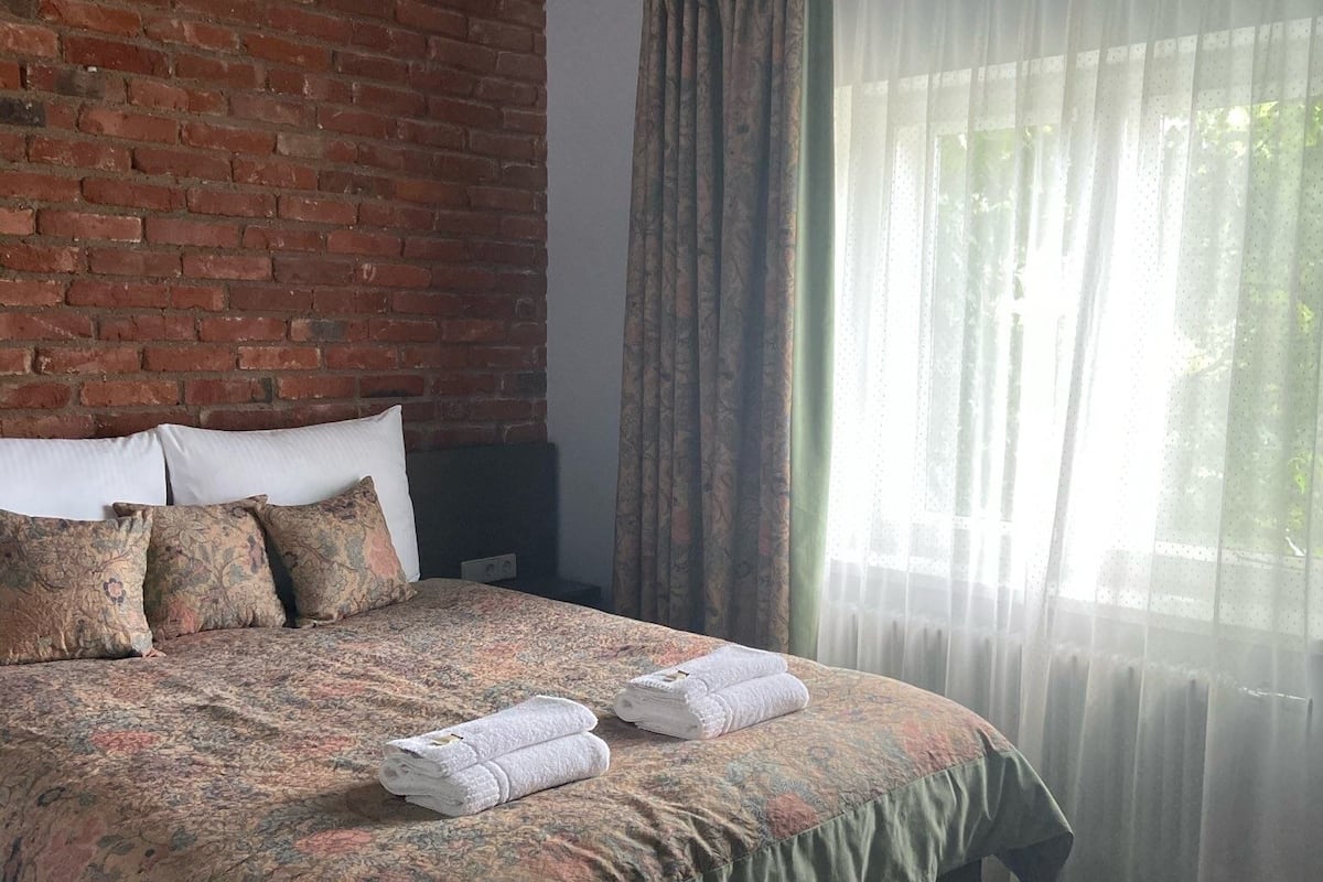 Une chambre avec un mur en briques, un lit recouvert d’une literie à motifs floraux, un tapis vert et une fenêtre ornée de rideaux légers.