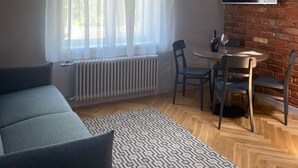 Een moderne woonkamer met een bank, een eettafel met stoelen, een tv die aan een bakstenen muur is bevestigd, en een raam met dunne gordijnen.