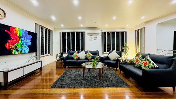 A modern living room with a flat-screen TV, black sofas, a coffee table, and a large window with blinds. - 3 Bedroom Apartment in Suva (Suva)