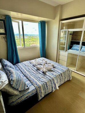 A neatly made bed with a patterned bedspread, a window with curtains, and a view of greenery outside. - Wonderful One Bedroom at Avida No. 40 (Iloilo)