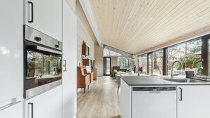 Eine moderne Küche mit weißen Schränken, einer Holzbalkendecke und großen Fenstern mit Blick ins Freie.