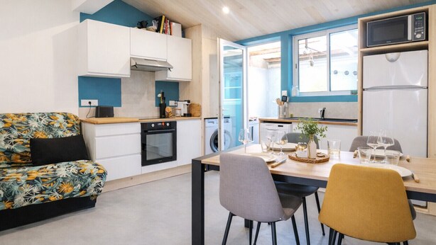 A compact kitchen with a microwave, oven, and washing machine. There is a dining area with a table set for two, and a sofa with a floral pattern.