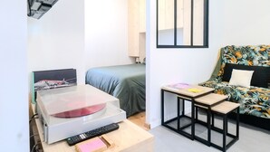 A compact room with a bed, a small table with a remote control, a decorative item, and a wall-mounted shelf.
