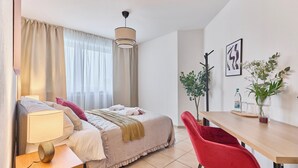 A hotel room with a bed, bedside table, lamp, red chair, dining table, potted plants, and artwork on the wall. - Rosalia - Stylish living, central experience (Kaiserslautern)