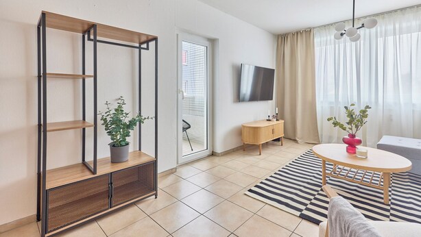 A modern living room with a wooden shelving unit, a round table, a TV mounted on the wall, and a large window with sheer curtains. - Rosalia - Stylish living, central experience (Kaiserslautern)