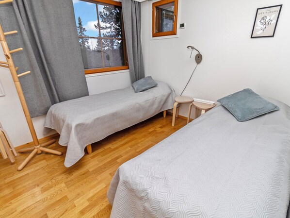A bedroom with two single beds, a wooden floor, a window with a view of trees, a bedside table with a lamp, and a wall-mounted power outlet.
