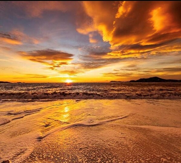 Une plage au coucher du soleil, avec des vagues et des montagnes à l’horizon.