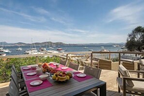 Mesa lista con platos, tazones y frutas, con vista a un puerto deportivo con barcos y colinas al fondo.