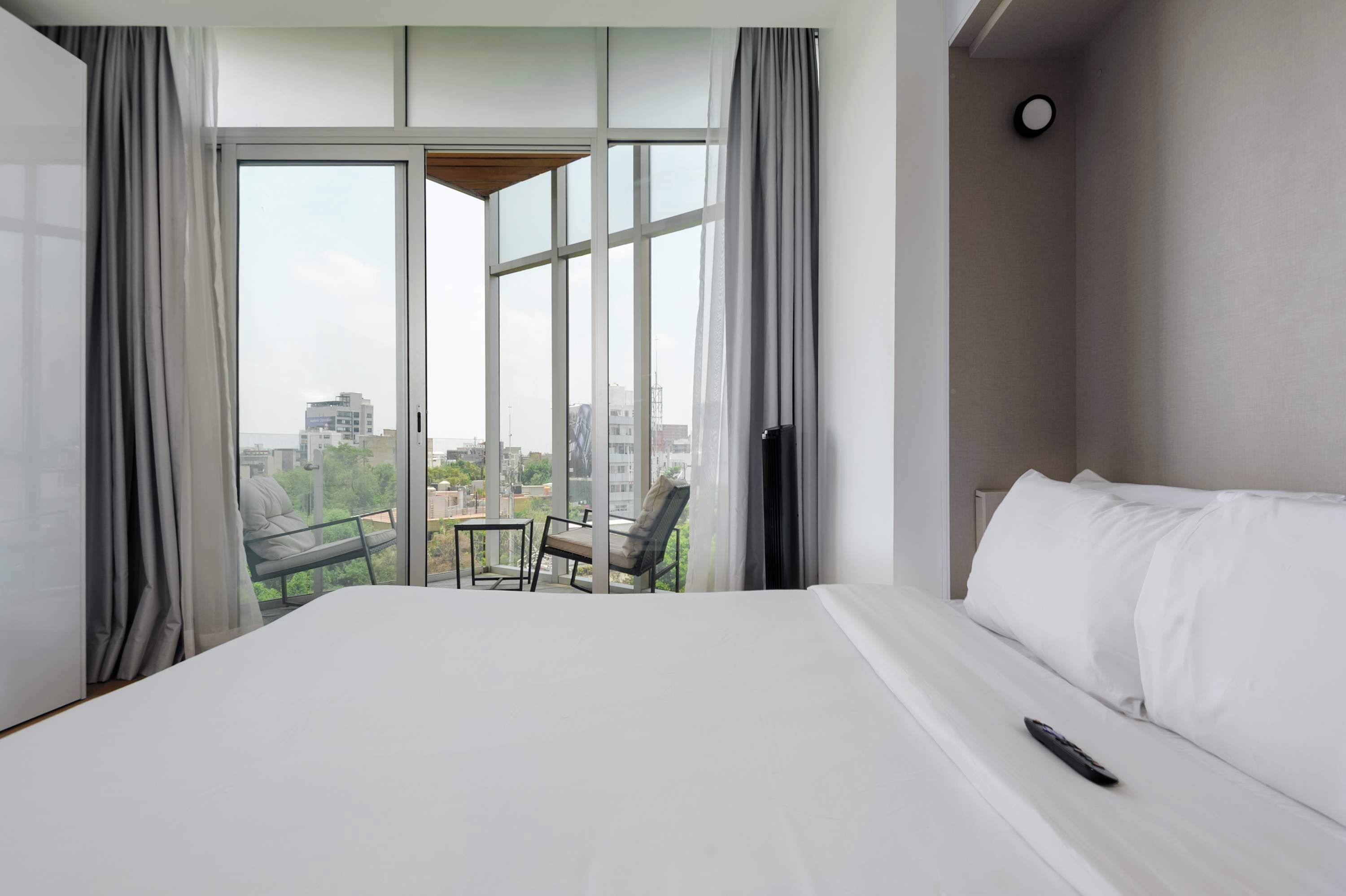 A hotel room with a large bed, a TV, a balcony with a table and chairs, and a view of the city.