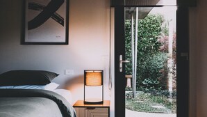 Quarto com cama, luminária de cabeceira, porta que dá para um jardim e uma obra de arte emoldurada na parede.