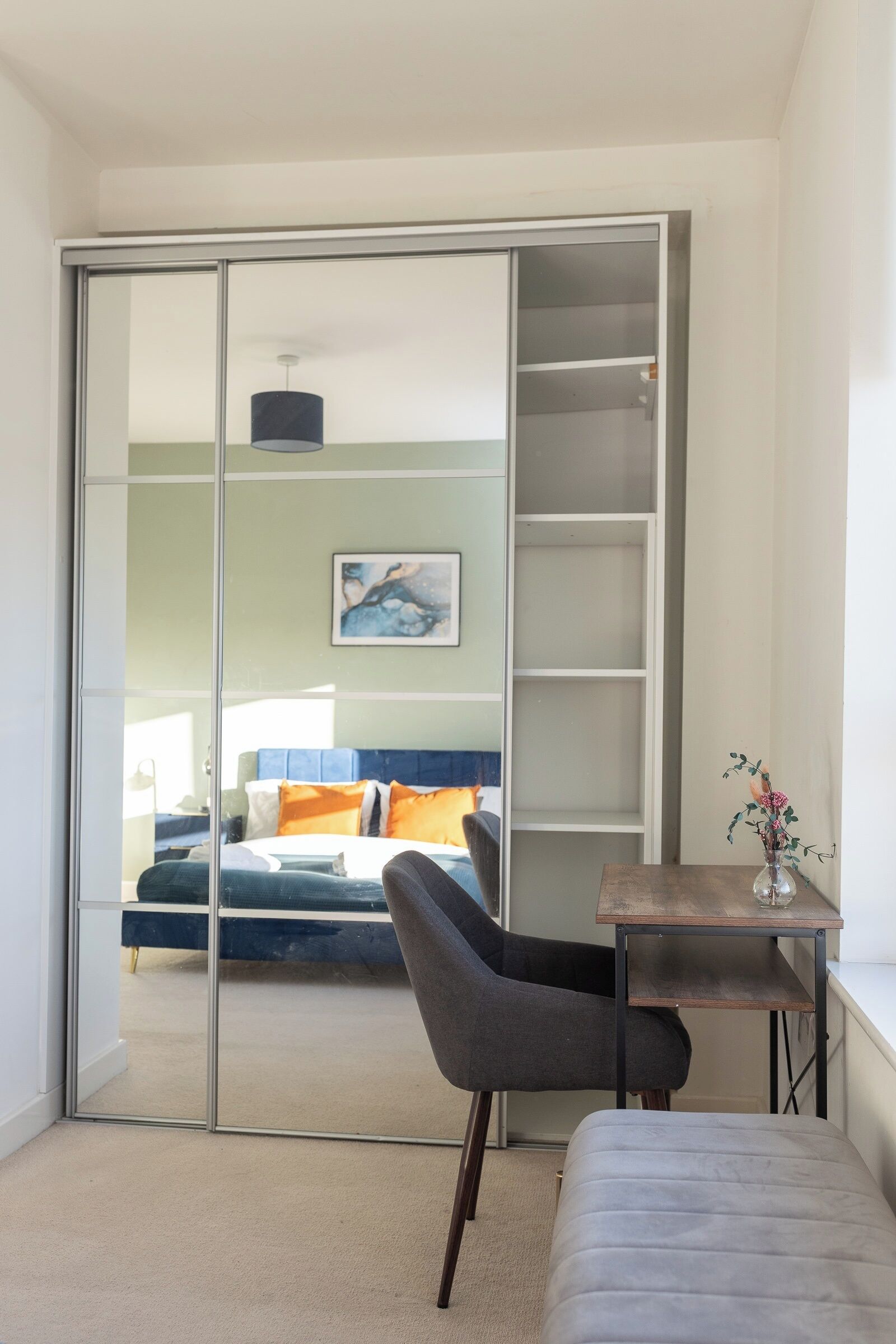 Een moderne slaapkamer met een schuifdeur naar een balkon, een grijze stoel, een klein houten tafeltje met een vaas en een bed met blauwe en oranje beddengoed.
