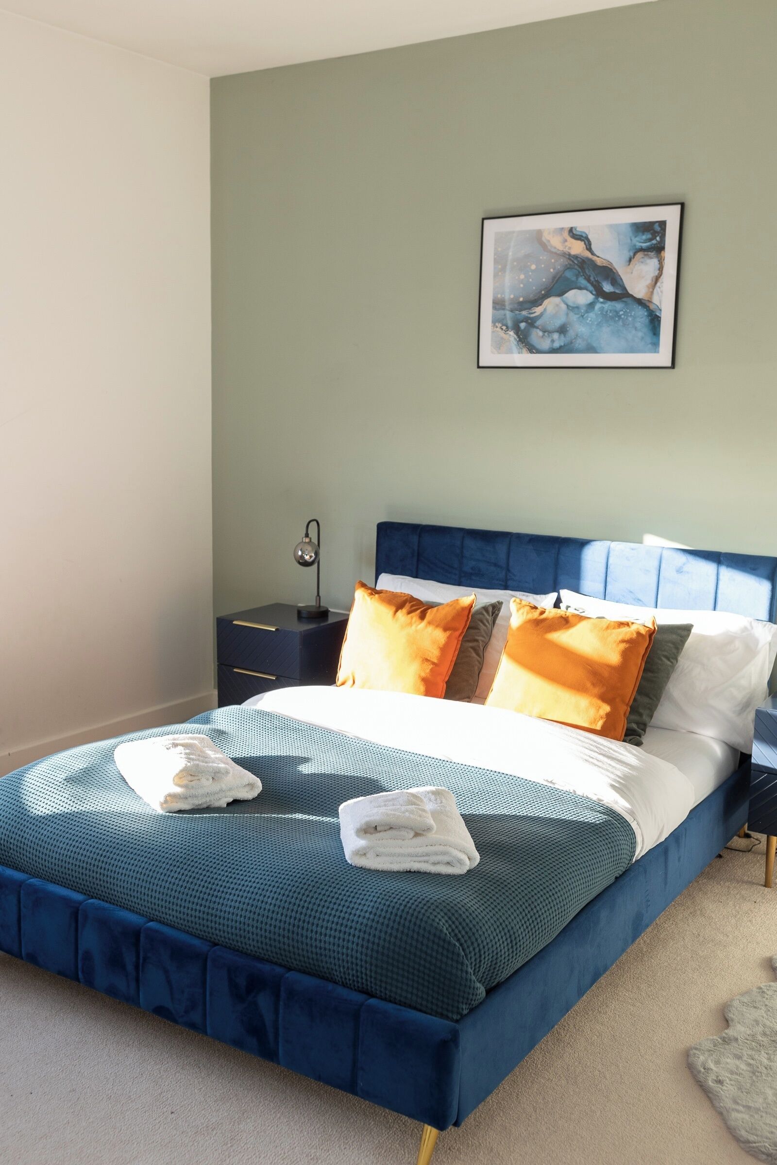 A neatly made bed with blue bedding and orange pillows, a bedside table with a lamp, and a framed abstract painting on the wall.
