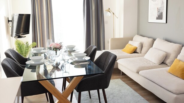 A modern living room with a glass dining table, black chairs, a beige sofa with yellow cushions, and a flat-screen TV mounted on the wall. - Stylish 2BR Apartment, Town Centre, Free Parking (Hatfield)